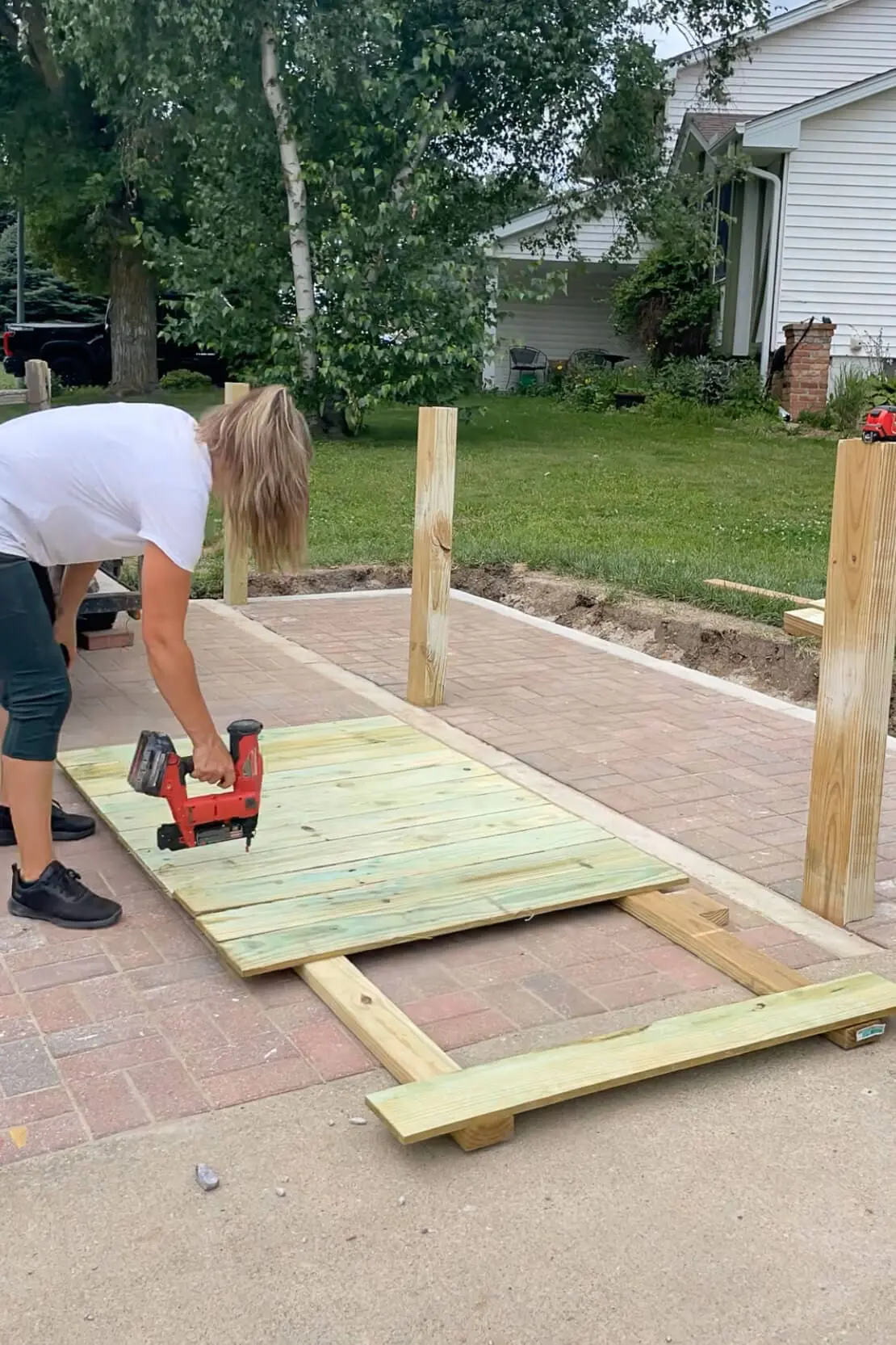 Woman using a nail gun to attach vertical fence pickets to a horizontal frame on a brick paver pad for a DIY trash enclosure panel.